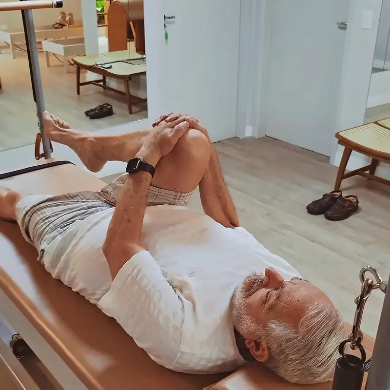 Aluno mais velho deitado no Reformer, em exercício acompanhado, em sala clara.